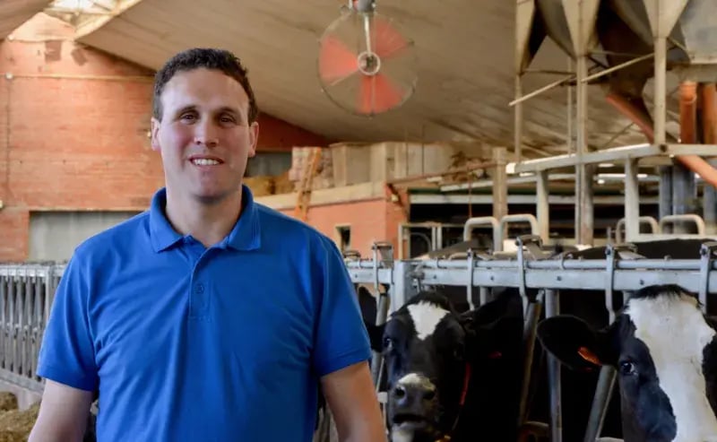 Un homme souriant, vêtu d'un polo bleu, se tient dans un bâtiment de Vache Laitière. Il est entouré de vaches à l'arrière-plan, avec un grand ventilateur visible au-dessus. Le bâtiment présente un haut plafond et des murs en briques, suggérant un environnement agricole bien entretenu.