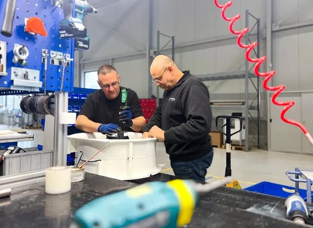 Zwei Männer arbeiten in einer Fabrik an der Montage eines Maschinenteils, mit Werkzeugen und Geräten um sie herum