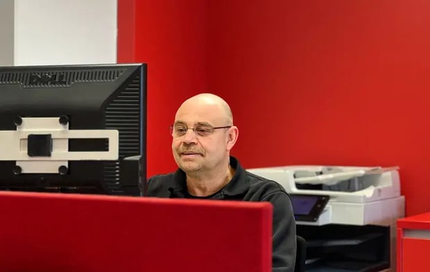 Mann arbeitet an einem Computer in einem Büro mit roter Wand und Drucker im Hintergrund