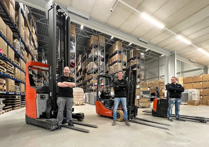 Drie magazijnmedewerkers poseren met heftrucks tussen hoge stellingen