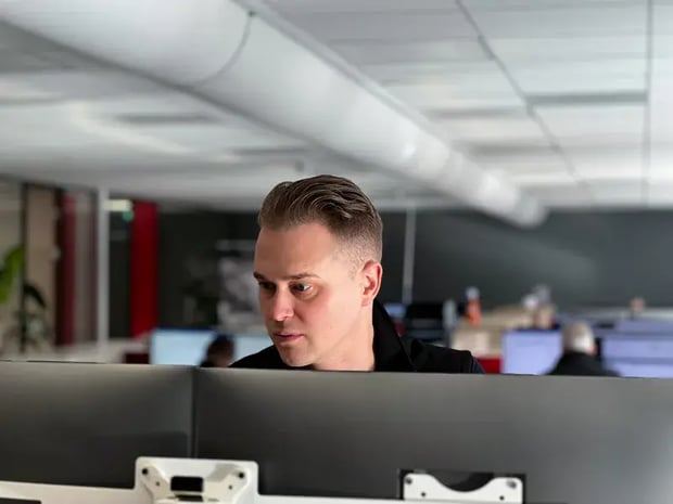 Ein Mann in einem Büro, der konzentriert auf zwei Computermonitore schaut. Im Hintergrund sind weitere Personen und Computerarbeitsplätze zu sehen.