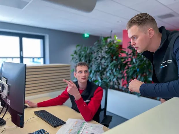 Zwei Männer in einem Büro, die gemeinsam an einem Computer arbeiten. Der sitzende Mann zeigt auf den Monitor, während der stehende Mann aufmerksam zuhört und nach vorne gebeugt ist.
