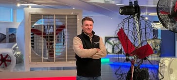 Un hombre está de pie con los brazos cruzados frente a grandes ventiladores industriales en una sala de exposición bien iluminada. El fondo muestra varios ventiladores y una iluminación colorida.