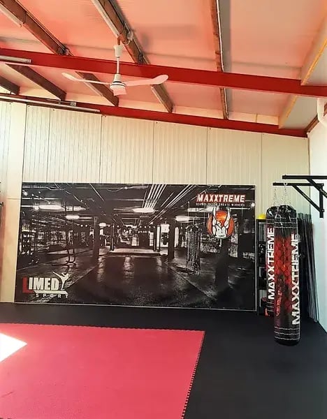 Vista interior de un gimnasio o espacio de entrenamiento con una combinación de colores rojo y negro. Del techo cuelga un ventilador que proporciona ventilación. La pared muestra un gran mural en blanco y negro que representa una escena de un gimnasio de boxeo, mientras que el suelo está ocupado por una colchoneta roja. También se ve un saco de pesas, rotulado con la marca «MAXXTREME».