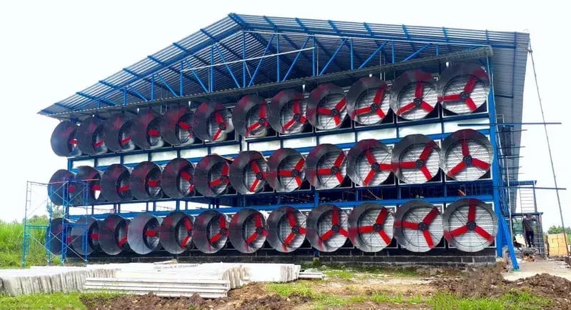 这是一栋大型工业建筑,外墙安装了许多大型通风扇。这些通风扇有防护格栅和红色叶片,并排成多排。建筑结构由蓝色金属框架支撑,部分被建筑材料包围,表明这是一项在建的建筑项目。