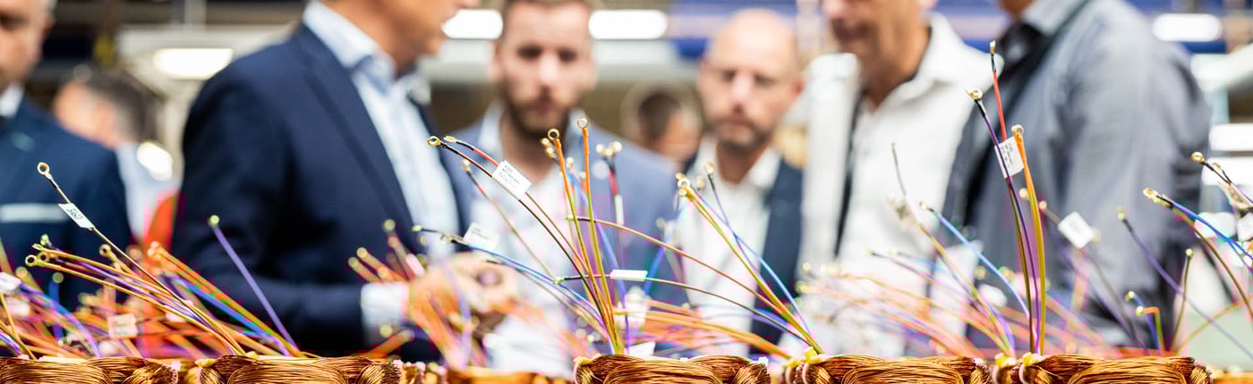 Een groep mannen in zakelijke kleding bekijkt een verzameling kleurrijke elektrische bedrading en koperen spoelen in een productiefaciliteit.