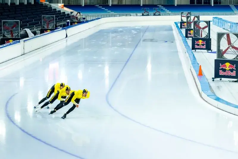 Patinadores de velocidad en trajes amarillos corriendo en una pista de hielo con grandes ventiladores en el fondo.